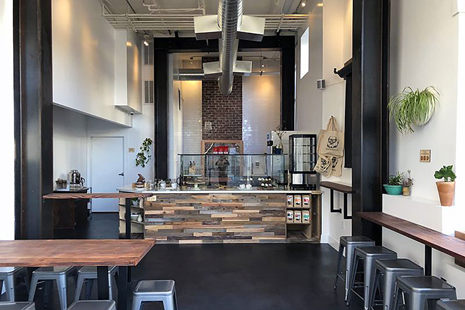 Bakery interior. Custom serving counter area built with reclaimed wood, stone countertops, and glass display racks. Space has high ceilings, white walls with hanging plants and display art. Polished concrete floor